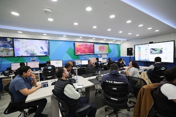 Sala de monitoreo con varias pantallas, mesas de trabajo, sillas, teclados de computadora, personas sentadas y el logo de MAGA en algunos monitores