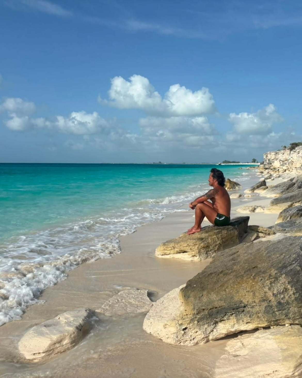 Torres, sin remera y con una malla negra corta, sentado sobre rocas en la orilla, con la vista fija en el horizonte y el mar abierto como único fondo
