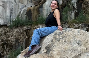 Mujer joven sentada en una roca grande con una camiseta negra sin mangas, jeans rasgados y zapatillas marrones. Fondo de rocas con vegetación y agua