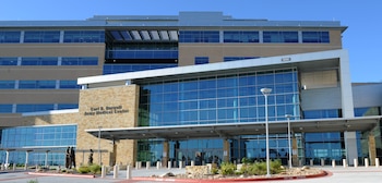 Fachada de un edificio moderno con ventanas de cristal azul, paredes de piedra, cartel "Carl R. Darnall Army Medical Center", estatuas y camino circular