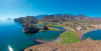Vista aérea de un paisaje costero con montañas áridas, un campo de golf verde, cuerpos de agua azul, una playa oscura y un complejo de edificios
