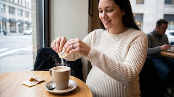 Una mujer embarazada sonriente vierte Splenda de un paquete amarillo en su café, sentada en una mesa de madera junto a una ventana con vista a una calle urbana.