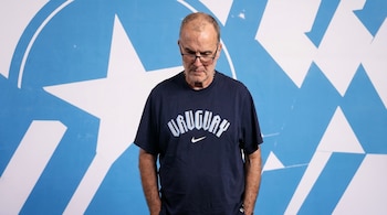 Un hombre de mediana edad, calvo, con gafas y una camiseta azul marino con la inscripción "URUGUAY", mira hacia abajo sobre un fondo azul con figuras blancas.