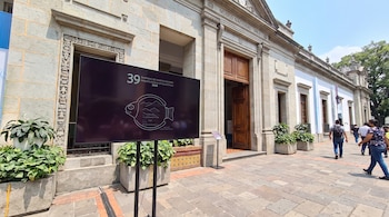 Exterior de un edificio de piedra con puerta de madera, frente al cual hay una pantalla negra con texto y un dibujo de pez, plantas en macetas y personas.