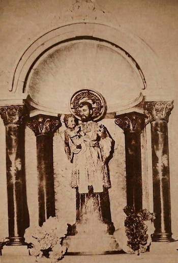 Escultura monocromática de San Cayetano que sostiene al Niño Jesús, ambos con halo. La escultura está en un nicho con dos columnas y decoraciones florales en la base