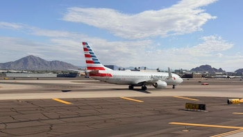 Un avión blanco de American Airlines con cola roja, blanca y azul en una pista de aeropuerto bajo un cielo azul, con montañas en la distancia