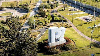 Vista aérea del Parque de la Innovación en Núñez con césped, árboles, senderos y una gran escultura blanca con las letras "PI". Se ven farolas y bancos