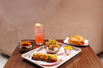 Una brocheta de carne, un cuenco con arroz salteado, salsas, una empanada y una bebida rosada con hielo sobre una mesa de madera