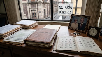Mesa de notaría con documentos, un libro "Escrituras", calendario con fechas, retrato de pareja, llaves, reloj. Cartel "Notaría Pública" visible por ventana.