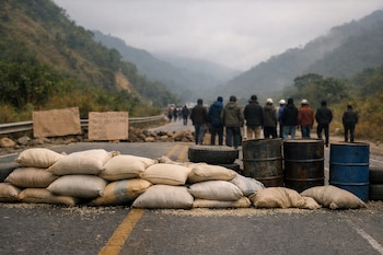 Vista de una carretera bloqueada en Perú con sacos de arroz, barriles y letreros improvisados; personas borrosas se ven al fondo con montañas cubiertas de niebla.