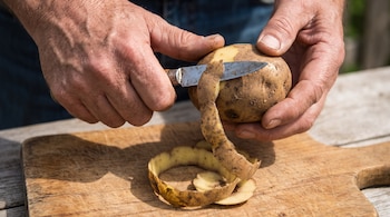 Dos manos sujetan y pelan una papa con un cuchillo. Una cáscara larga y varias pequeñas están sobre una tabla de madera.