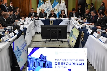 Personas en traje formal y uniformes oficiales están sentadas alrededor de una mesa en forma de herradura con micrófonos y pantallas