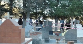 Personas detrás de una cinta de seguridad entre lápidas de piedra, cruces y árboles en un cementerio