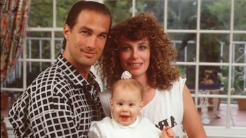 Steven Seagal, Kelly LeBrock y un bebé en primer plano. Seagal lleva una camisa a cuadros, LeBrock tiene cabello rizado. Fondo de ventana grande