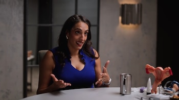La Dra. Rena Malik, una mujer con cabello oscuro y vestido azul, sentada a una mesa blanca, gesticula mientras habla. Un modelo de útero está a su derecha