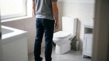 Un hombre visto de espaldas con camiseta gris y pantalón oscuro está de pie frente a un inodoro blanco en un cuarto de baño iluminado.