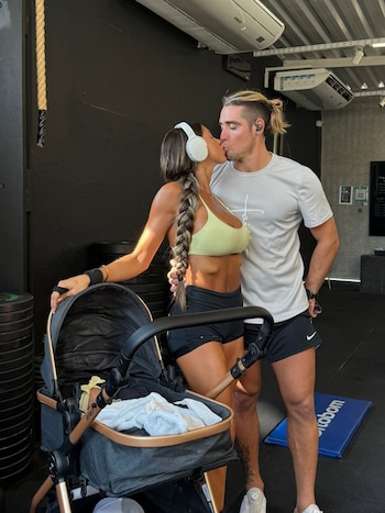 Una mujer y un hombre con ropa deportiva se besan en un gimnasio junto a un cochecito de bebé