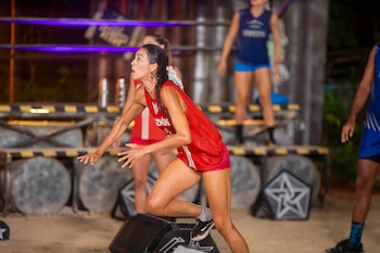 Atleta femenina en un uniforme rojo, mojada y en posición dinámica de carrera en un circuito de obstáculos con iluminación púrpura de fondo