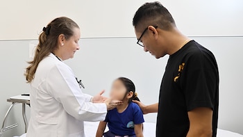 Una médica con bata blanca y estetoscopio palpa el cuello de una niña sentada, junto a un hombre con gafas que la sostiene del hombro