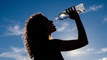 Silueta de una mujer bebiendo agua directamente de una botella de plástico. El sol brilla por detrás de ella contra el cielo azul con nubes dispersas.