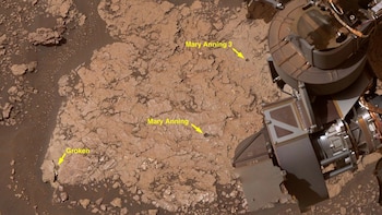 Vista aérea de la superficie marciana con rocas rojizas fracturadas y arena. Una parte del equipo del rover, posiblemente Mastcam, es visible a la derecha. Flechas amarillas indican tres perforaciones en la roca