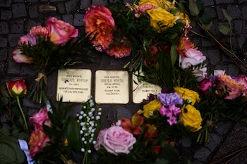 Primer plano de tres adoquines Stolpersteine dorados en un camino de piedra, rodeados por múltiples ramos de flores frescas de colores vivos