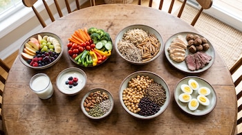 Ocho platos y boles con frutas, verduras, cereales, carnes, legumbres, frutos secos, huevos y lácteos sobre una mesa de madera.