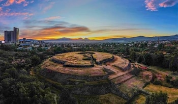 Vista aérea de una pirámide circular escalonada rodeada de vegetación, con edificios urbanos y montañas en el horizonte bajo un cielo anaranjado y azul