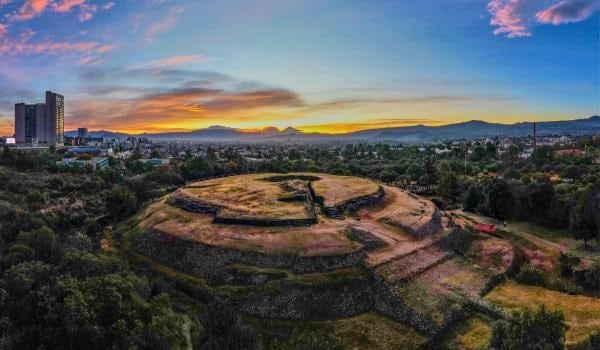 La Pirámide de Cuicuilco, una estructura prehispánica, se alza en la zona arqueológica con edificios modernos y montañas en el horizonte bajo un cielo de tonos cálidos. (INAH/Cuicuilco)