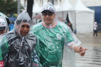Dos hombres usando ponchos de lluvia, uno con camiseta verde de México y un visor blanco, el otro con poncho negro, con gotas de lluvia y carpas blancas al fondo