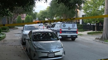 Cinta amarilla de la policía bloquea una calle residencial con un coche plateado Toyota en primer plano y un vehículo de la Unidad de Escena del Crimen al fondo