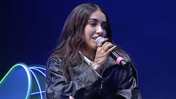 Maria Becerra, con cabello oscuro y chaqueta de cuero negra, sostiene un micrófono frente a un fondo azul oscuro con detalles de neón