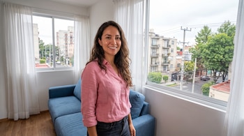 Mujer con blusa rosa de pie junto a ventana, con sofá azul detrás. Se ven cortinas blancas y un barrio de Ciudad de México con edificios y árboles.