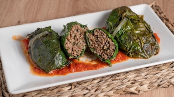 Rollos de hojas de acelga rellenos de carne picada, uno cortado a la mitad, sobre salsa de tomate en un plato blanco.