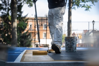 Un trabajador con pantalón manchado de pintura utiliza un rodillo para pintar el piso de una cancha deportiva al aire libre