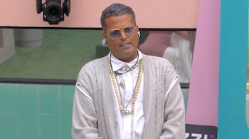 Aldo Rendón viste un cárdigan gris, camisa blanca, collares y gafas de cristales amarillos frente a un fondo de color rosa y verde.