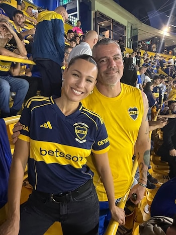 Pampita sonríe junto a un hombre en un estadio de fútbol. Ambos visten camisetas de Boca Juniors. Al fondo se observan gradas llenas de aficionados