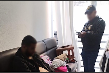 Un hombre de perfil sentado en un sofá oscuro con la cara pixelada, junto a ropa y una cesta. Un oficial de policía con gorra y chaleco revisa un teléfono