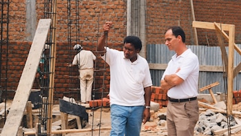 Dos hombres conversan en una obra en construcción frente a columnas de varillas de hierro y muros de ladrillo, con un obrero al fondo