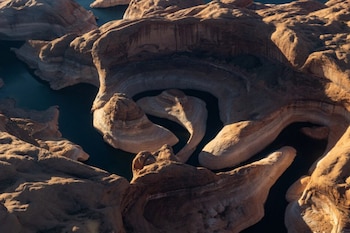 Vista aérea del Río Colorado oscuro serpenteando entre formaciones rocosas sedimentarias claras, mostrando capas horizontales y sombras contrastantes