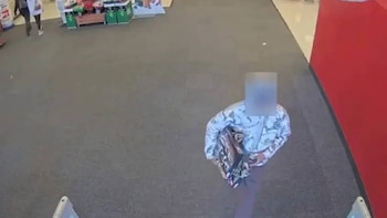 Vista aérea de una persona con rostro borroso y ropa estampada, de pie en un piso gris de un establecimiento. Se aprecian estantes verdes y una pared roja