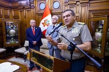 Un hombre uniformado con muchas medallas habla en un atril ante un micrófono, con la bandera de Perú y dos hombres de traje en un salón con paneles de madera