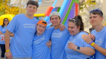 Cinco jóvenes sonrientes con camisetas azules 'Walking 4 Friendship' posan juntos al aire libre. Uno lleva gorra naranja, otro gorra beige. Se ven pulseras de colores y una botella de agua
