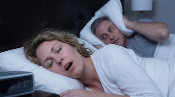 Una mujer duerme en la cama junto a un reloj digital mientras un hombre a su lado se tapa las orejas con una almohada.
