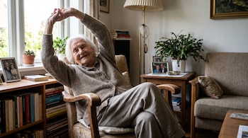 Mujer anciana de cabello blanco, sentada en un sillón, estirando los brazos hacia arriba y sonriendo. Visible una ventana, plantas y estanterías en el salón.