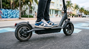 Plano cercano de los pies de una persona sobre la plataforma y las ruedas de un patinete eléctrico negro en movimiento sobre el asfalto.