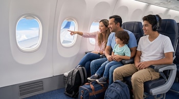 Una mujer, un hombre, un niño y un adolescente están sentados en un avión; la mujer señala la ventanilla y hay mochilas en el suelo.