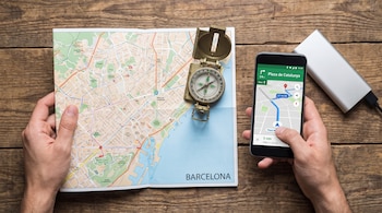 Una mano sostiene un mapa de papel de Barcelona, junto a una brújula y un smartphone con navegación GPS. Un cargador portátil se encuentra al lado.