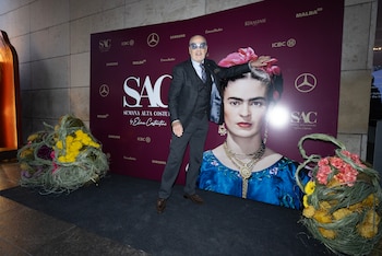 Un hombre con traje y gafas posa de pie junto a un cartel publicitario con un retrato de Frida Kahlo y arreglos florales en el suelo