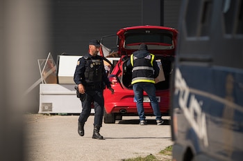 Un agente de policía en uniforme camina hacia la izquierda mientras otro, con chaleco reflectante, carga un paquete en el maletero abierto de un coche rojo